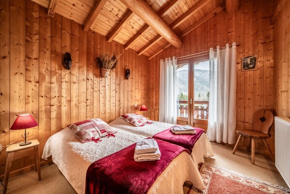 A wooden cabin room with two beds, a chair, a table, and a window with curtains.