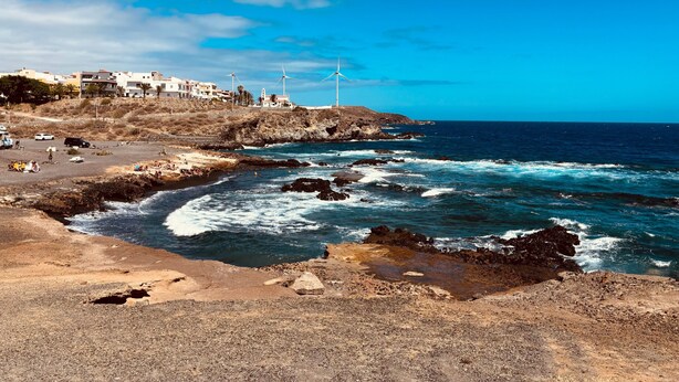 Uma área costeira com terreno rochoso, uma praia e edifícios com turbinas eólicas ao fundo.