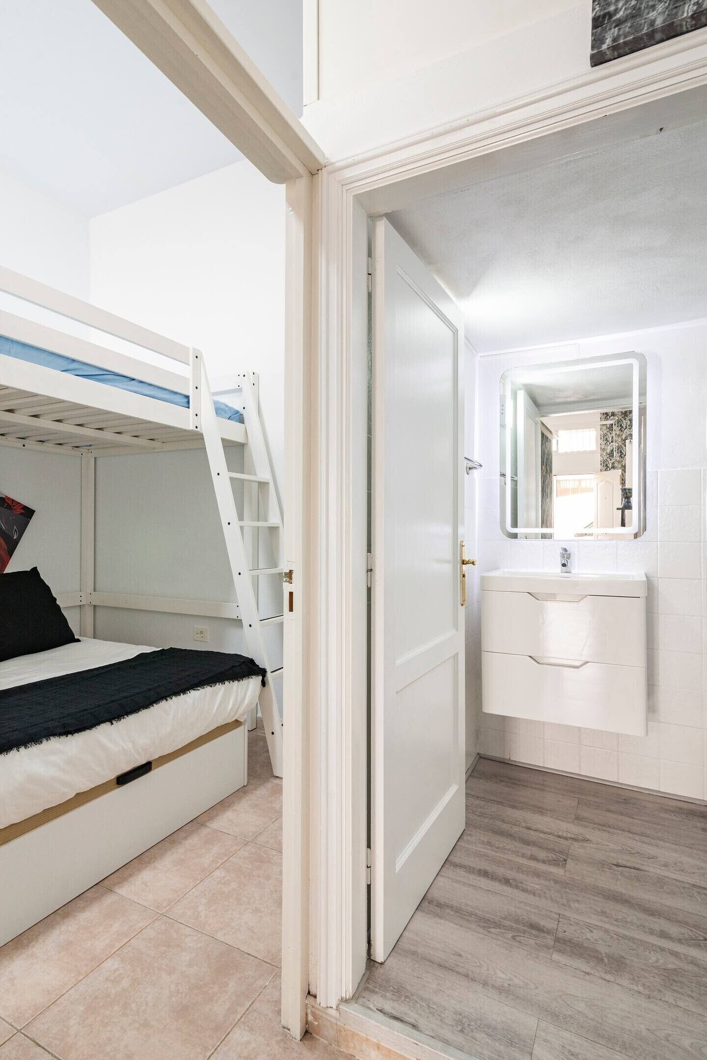 Una cama litera con escalera, un baño blanco con lavamanos y espejo, y suelo de madera.