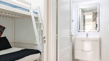 Una cama litera con escalera, un baño blanco con lavamanos y espejo, y suelo de madera.