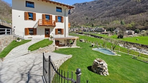 Une maison à deux étages avec une façade en pierre, un balcon et une pelouse bien entretenue agrémentée d’un petit étang.