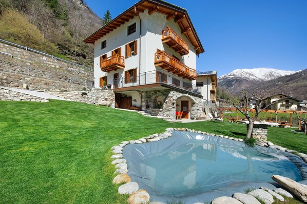 Un edificio de dos pisos con balcones, una escalera de piedra, piscina y montañas al fondo.