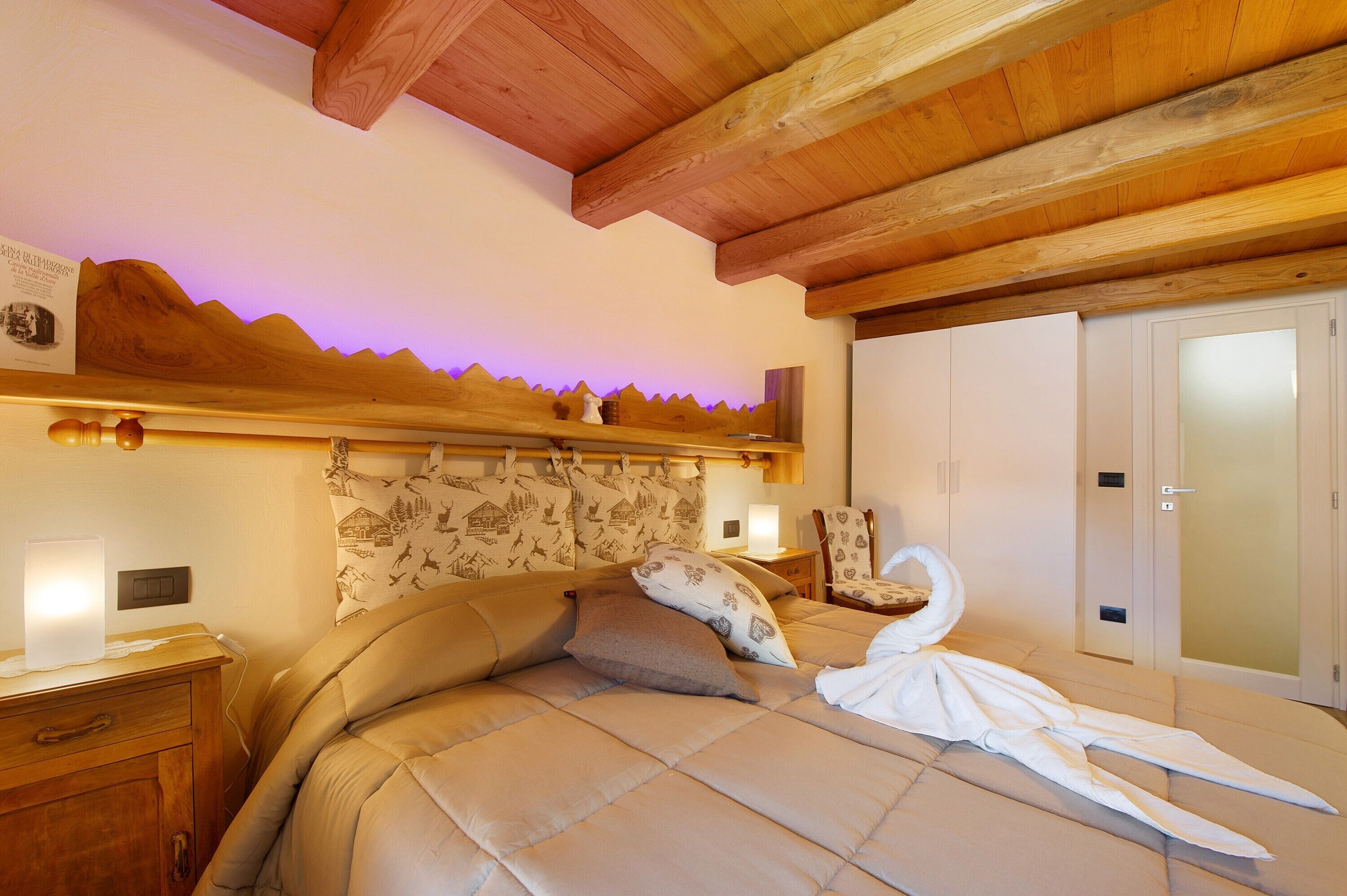Een slaapkamer met een houten plafond, een bed met een handdoek in de vorm van een zwaan, een nachtkastje, een stoel en een garderobe.