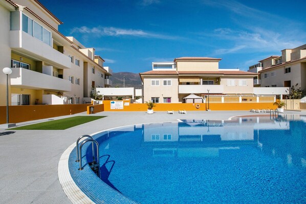 A swimming pool with a clear blue water, surrounded by buildings with balconies and a mountain in the background.