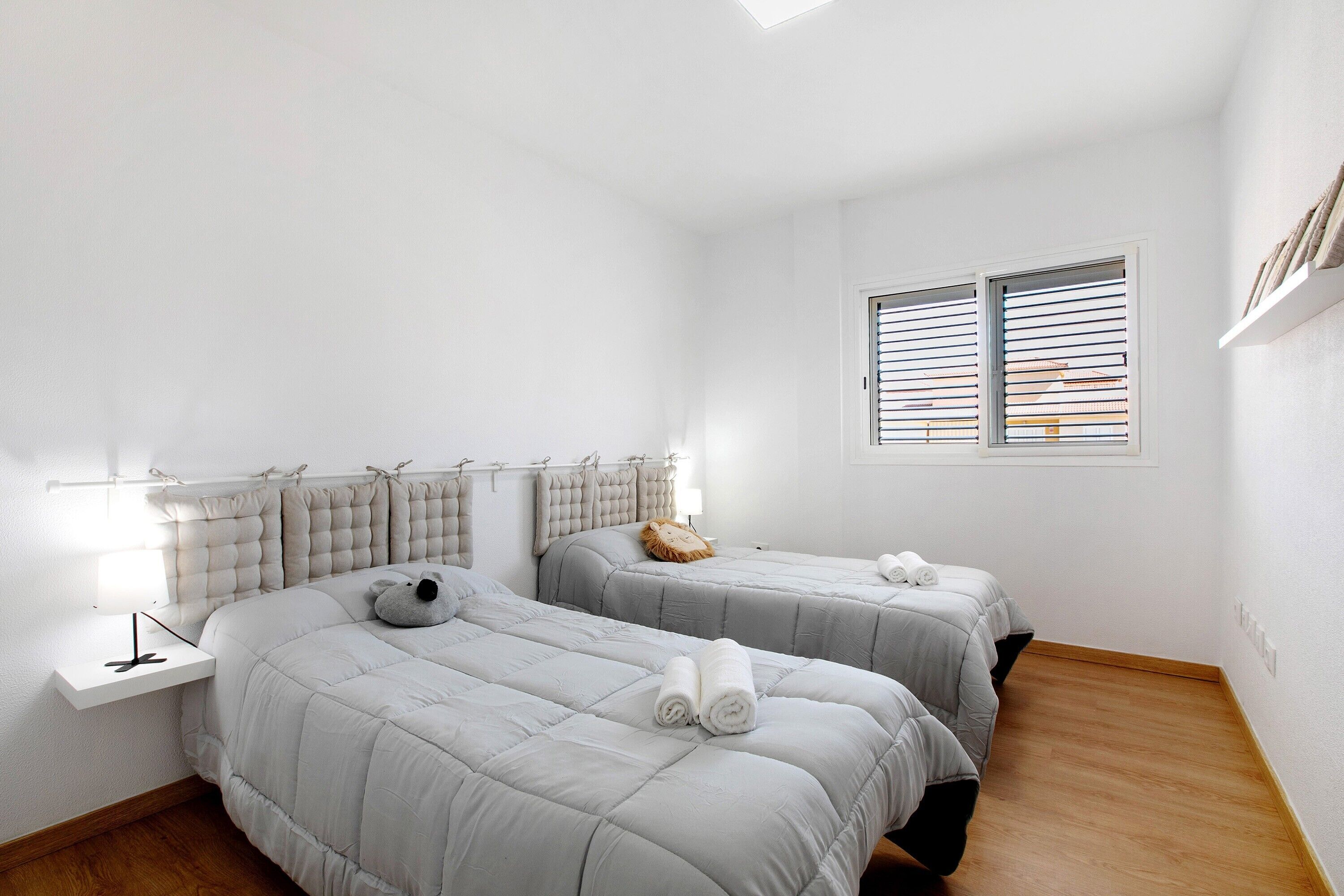 Una habitación con dos camas, una ventana con persianas, una mesita de noche con lámpara y un cabecero con diseño acolchado.