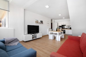 Ein modernes Wohnzimmer mit Flachbildfernseher, einem roten Sofa, einem blauen Sofa, einem weißen Esstisch und einer Küche im Hintergrund.