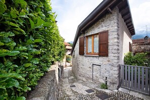 Une maison en pierre avec une fenêtre et des volets en bois, entourée de verdure et d’un mur de pierre.