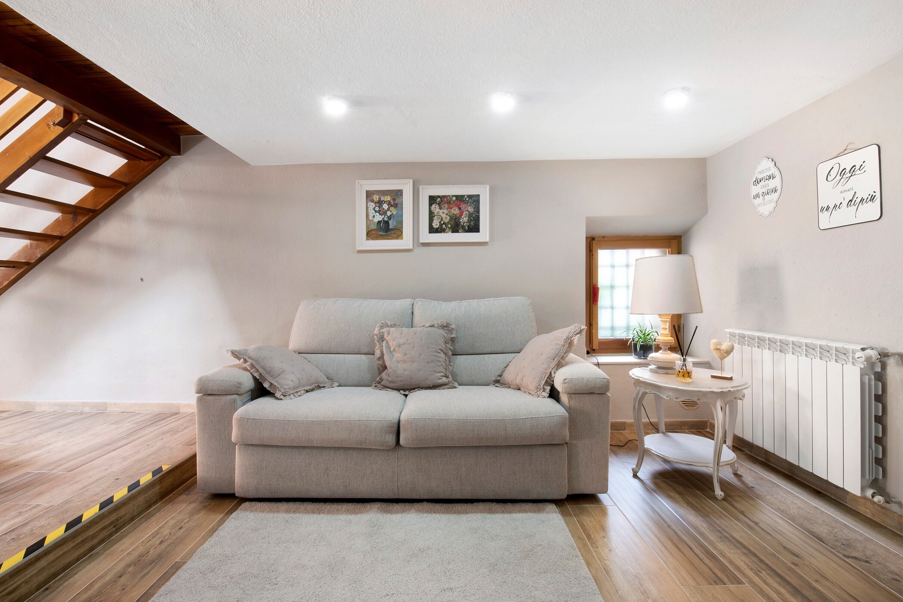 A living room with a grey sofa, a small round table with a lamp, and two framed pictures on the wall.