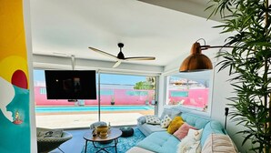 A living room with a sofa, a TV, a ceiling fan, and a view of a swimming pool. - Vacation Home "Casa Color" with Sea View, Private Pool & Wi-Fi (Corralejo)