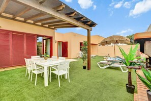 A patio with a white table and chairs, a thatched umbrella, and a small potted plant.