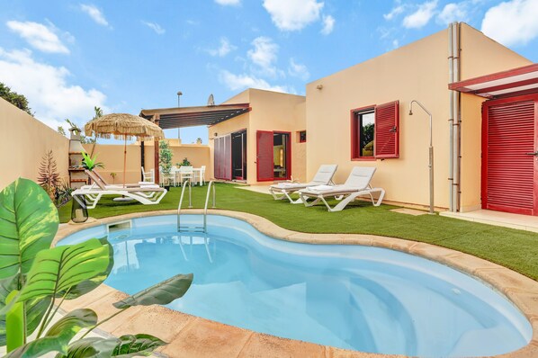 A swimming pool with lounge chairs, a covered patio, and a building with red shutters.