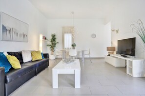 A modern living room with a sofa, coffee table, TV stand, dining area, and decorative plants.