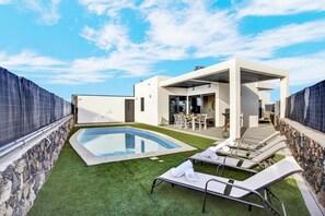 A modern outdoor pool area with a clear blue sky, a stone wall, and a white building with a covered patio.