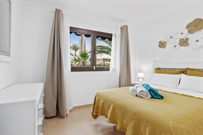 Quarto de hotel com uma cama, janela com cortinas, mesa de cabeceira com abajur e vista para palmeiras lá fora.
