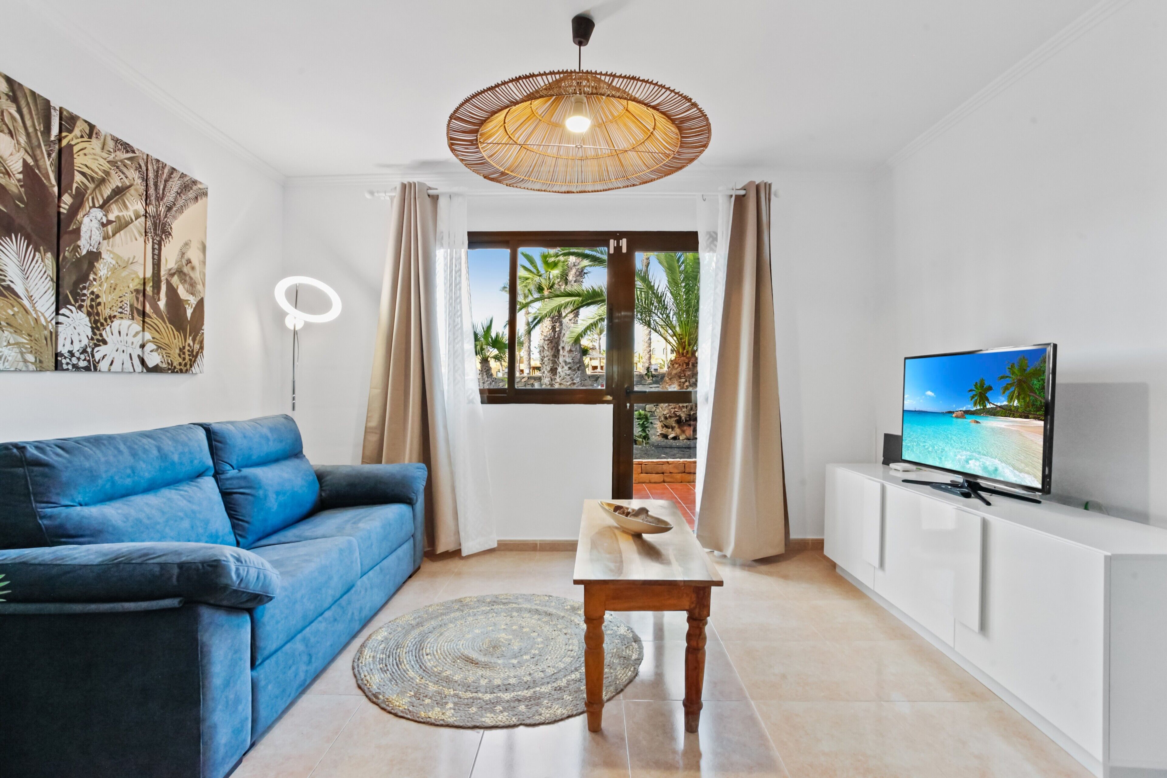 A living room with a sofa, a television, a table, a lamp, and a large window with a view of palm trees.