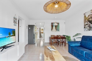 A modern living room with a flat-screen TV, a dining area with wooden chairs, a blue sofa, and a large painting on the wall.