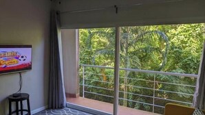 Sala de estar con televisor grande en la pared, sofá, un taburete pequeño y balcón con vista a zonas verdes.
