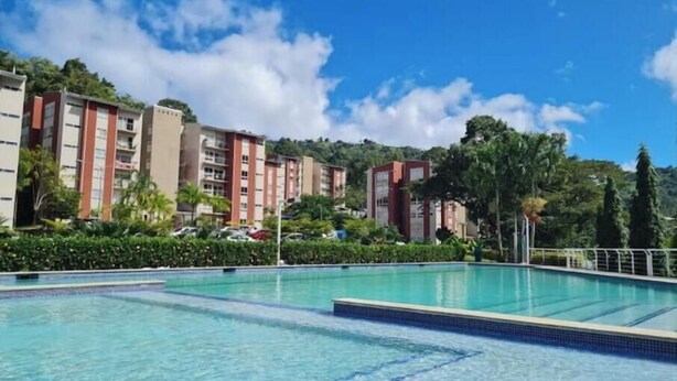 Una piscina con agua azul cristalina, rodeada por una valla. Al fondo, edificios de varios pisos con balcones y un paisaje montañoso con árboles.