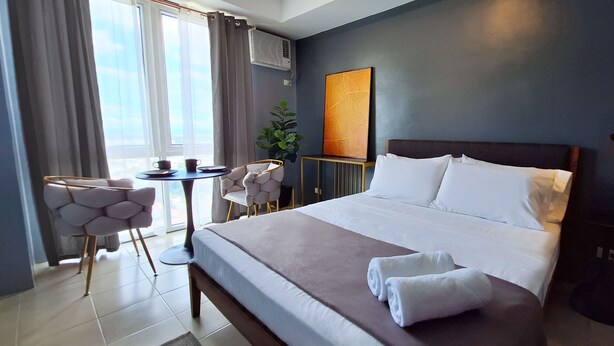 A modern hotel room with a large bed, a small round table with chairs, a potted plant, and a view of the outdoors through the window. - ELUDE Designer Suite Pasig (Prime location) (pasig city)