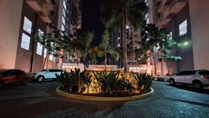 A nighttime view of a residential building with multiple floors, a landscaped garden area with palm trees, and parked cars. - ELUDE Designer Suite Pasig (Prime location) (pasig city)
