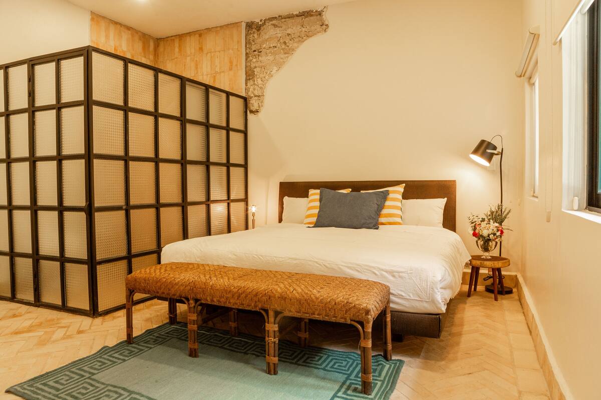 Un dormitorio con una cama grande, un banco, una mesita con una lámpara y un jarrón decorativo.