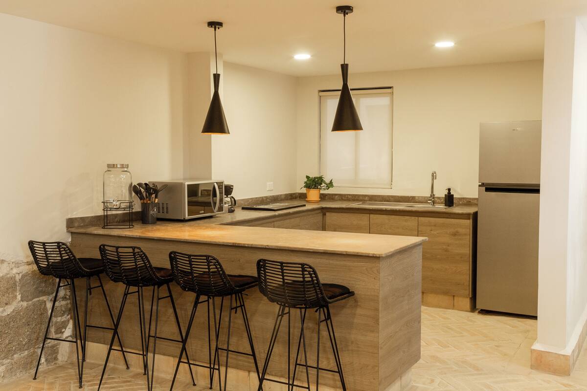 Una cocina moderna con una isla central, taburetes de bar, iluminación colgante, un refrigerador y un microondas.