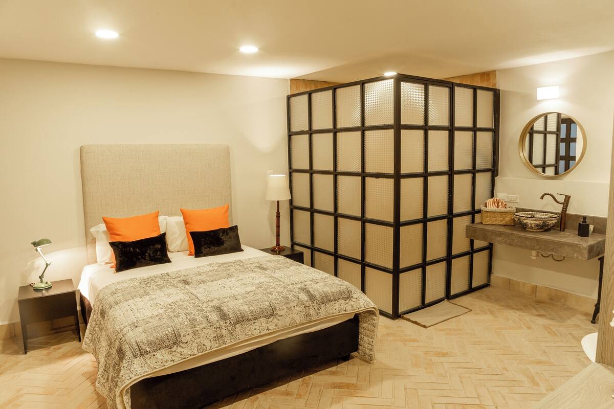 Un dormitorio con una cama grande, una mesita de noche, un tocador, un espejo, un panel de madera y un baño con lavamanos y espejo.