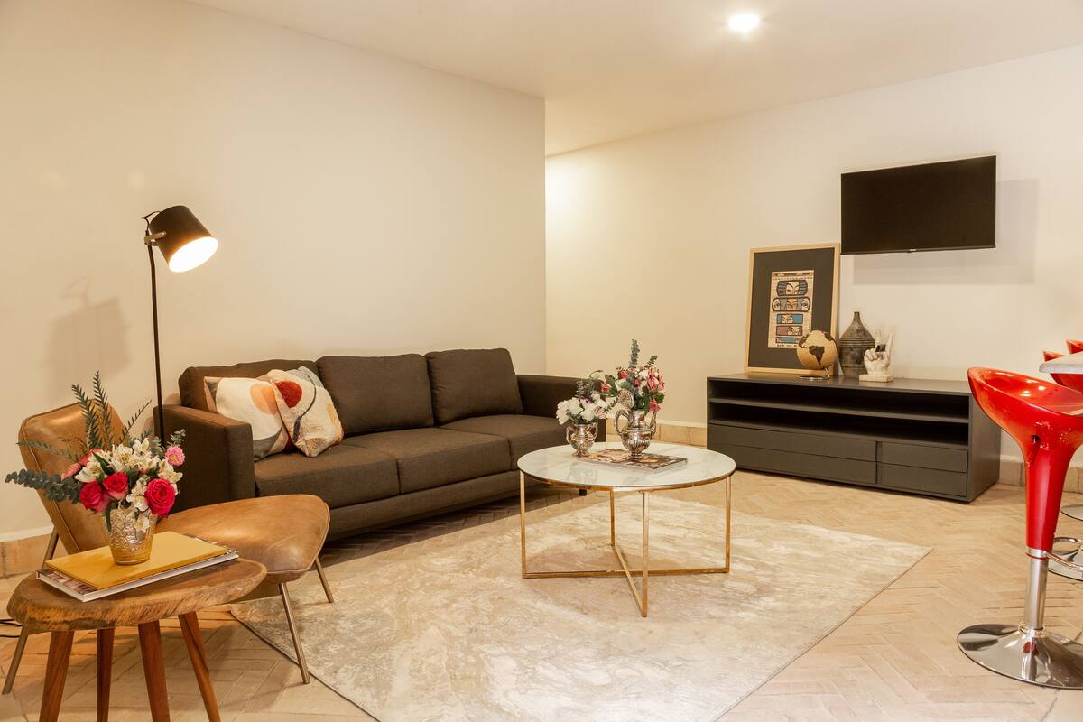 Una sala de estar moderna con un sofá, una mesa de centro, un taburete rojo, una lámpara de pie y un televisor de pantalla plana montado en la pared.