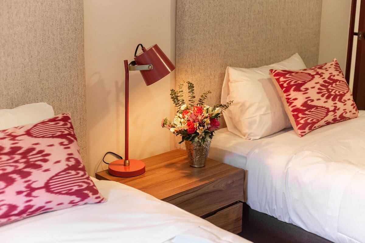 Habitación de hotel con dos camas, una mesita de noche con una lámpara y un jarrón con flores.