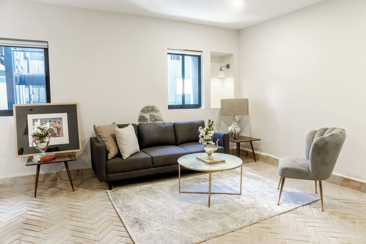 Una sala de estar moderna con un sofá, una mesa redonda para café, un sillón gris y un cuadro en un atril.