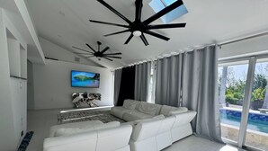 Een moderne woonkamer met een witte bank, plafondventilatoren, een groot raam met gordijnen, en een zwembad zichtbaar door de glazen deuren.