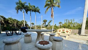 Een terras met een stenen tafel en banken, omgeven door palmbomen en een houten omheining.