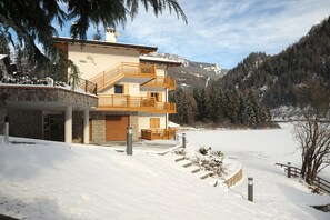 Un bâtiment à deux étages, doté d’une façade en pierre, de balcons en bois et offrant une vue sur un paysage enneigé avec des montagnes en arrière-plan.