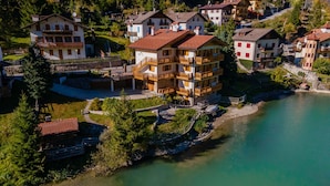A multi-story building with balconies, situated by a river with a bridge leading to a dock. - Villa Edelweiss Botton d'oro (Alleghe)