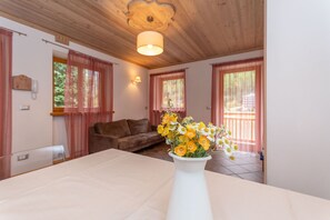 Une pièce avec un plafond en bois, un canapé marron, une table blanche ornée d’un vase de fleurs et une fenêtre avec des rideaux.
