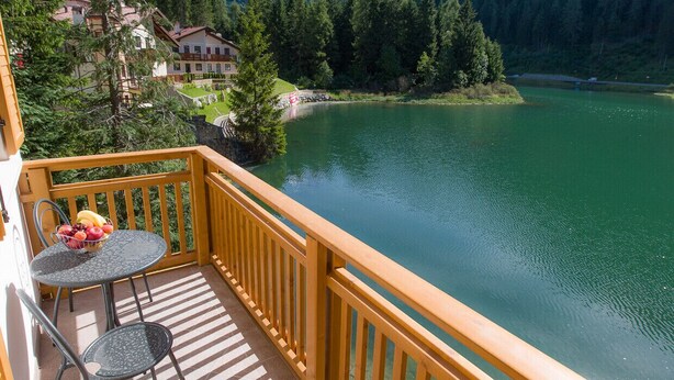 A balcony with a table and chairs overlooking a lake, surrounded by trees and buildings. - Villa Edelweiss Botton d'oro (Alleghe)