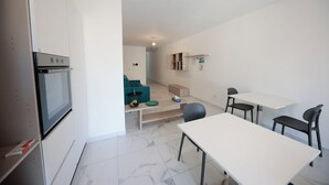 Una cocina moderna con horno empotrado, un comedor con mesa blanca y sillas negras, y una zona de estar con sofá verde y una mesa pequeña.