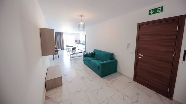 Habitación de hotel moderna con puerta de madera, sofá azul turquesa, suelo de mármol y una zona de cocina visible al fondo.