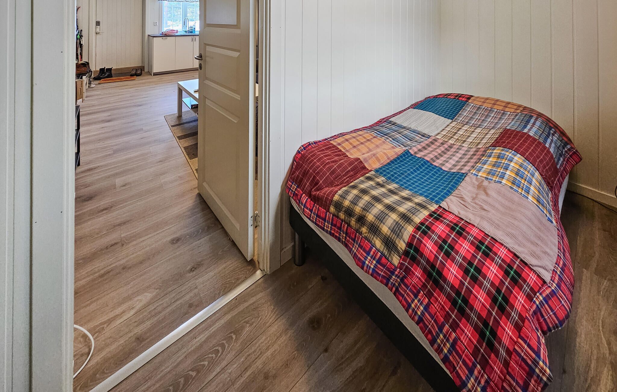 Un dormitorio con piso de madera, una cama con cubrecama a cuadros y una puerta blanca que da a una zona de cocina.