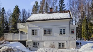 Ein zweigeschossiges weißes Holzhaus mit Balkon, umgeben von Schnee und Bäumen.