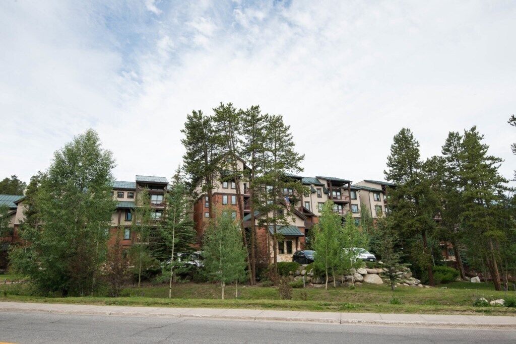 A multi-story residential building with a mix of brick and wood exteriors, surrounded by trees and a clear sky.