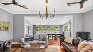 A spacious living room with a grey sofa, a brown leather couch, and a dining area with a table and chairs. A chandelier hangs from the ceiling, and there are ceiling fans on either side. - Prepare to Fall in Love (Millfield)