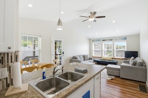 A modern kitchen with a double sink, a dining area with chairs, a grey sofa, and a ceiling fan.