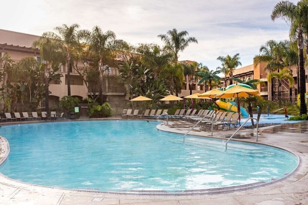 Un área de piscina con sillones de descanso y sombrillas, rodeada de palmeras y un edificio al fondo.