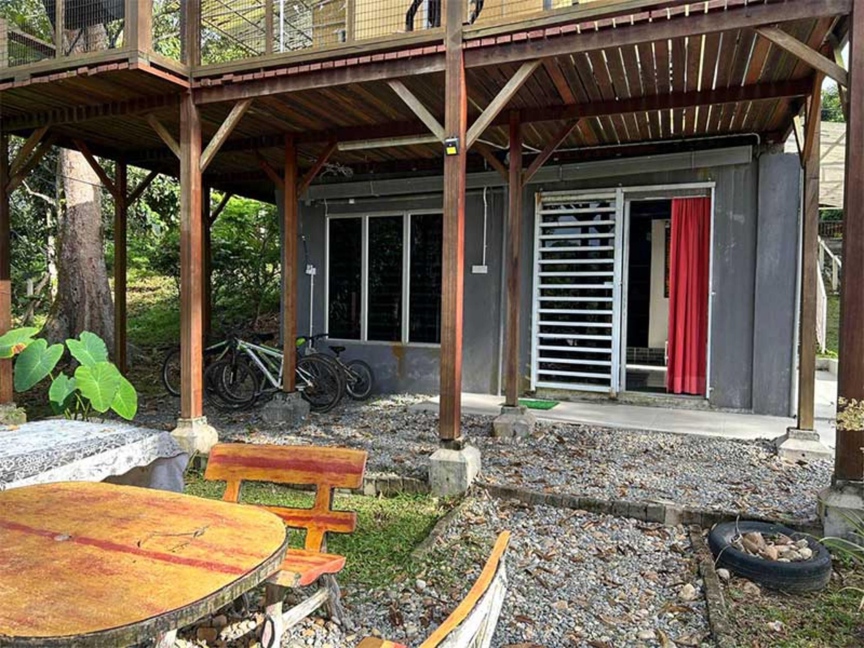 Un espacio al aire libre cubierto con un banco de madera, una mesa y una bicicleta estacionada bajo una estructura con baranda metálica y cortina roja.
