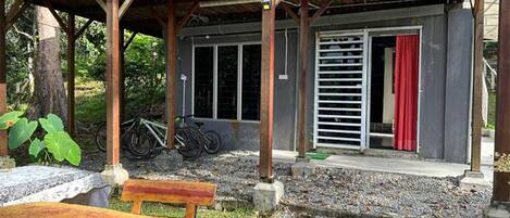 A covered outdoor area with a wooden bench, a table, and a bicycle parked under a structure with a metal railing and a red curtain.