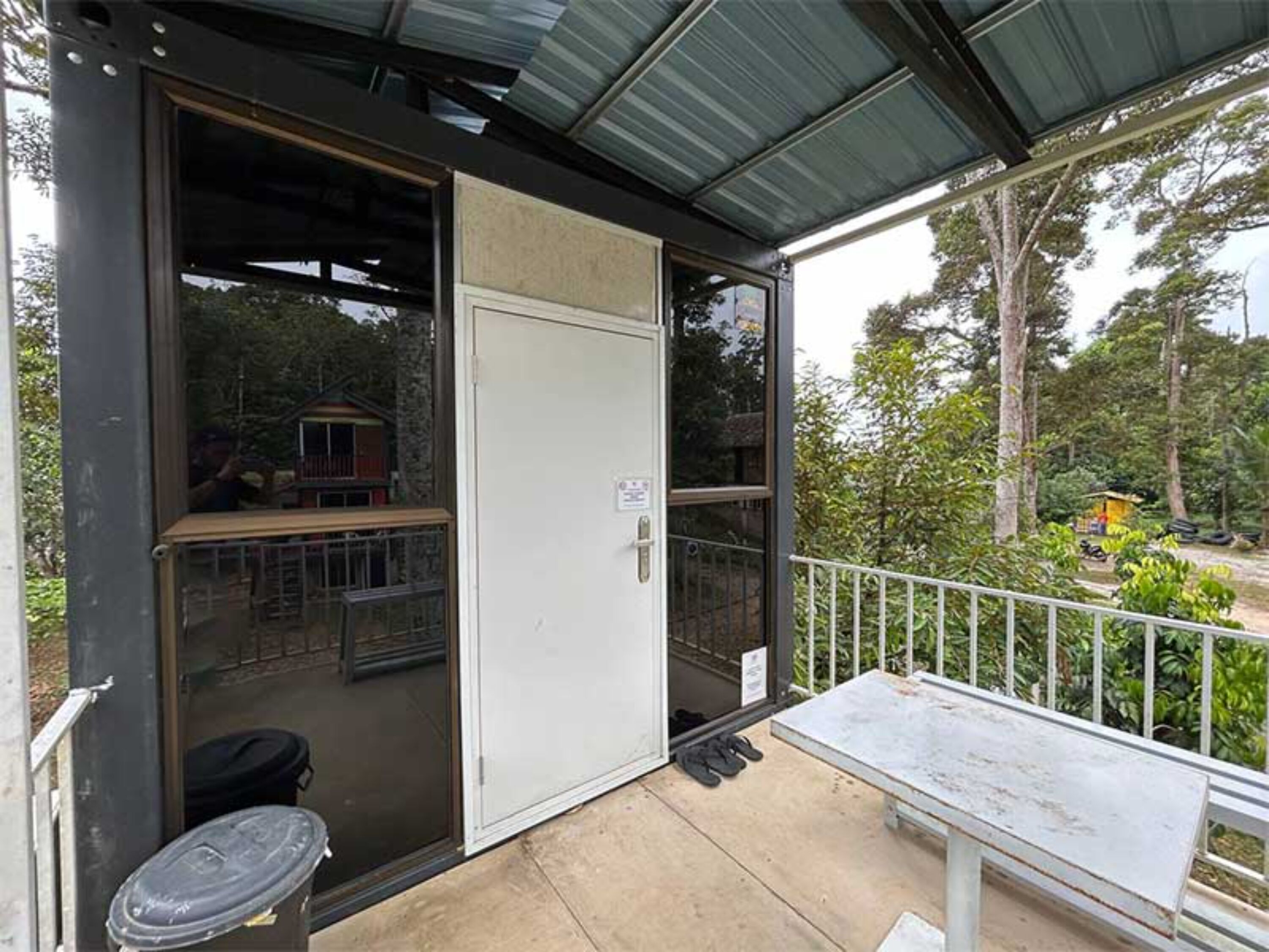 Un espacio al aire libre techado con una puerta corrediza de vidrio que da a una habitación, una pequeña mesa y un par de zapatos en el suelo.