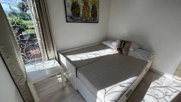 A bedroom with a bed, a window with curtains, a painting of pineapples, and a view of greenery outside.