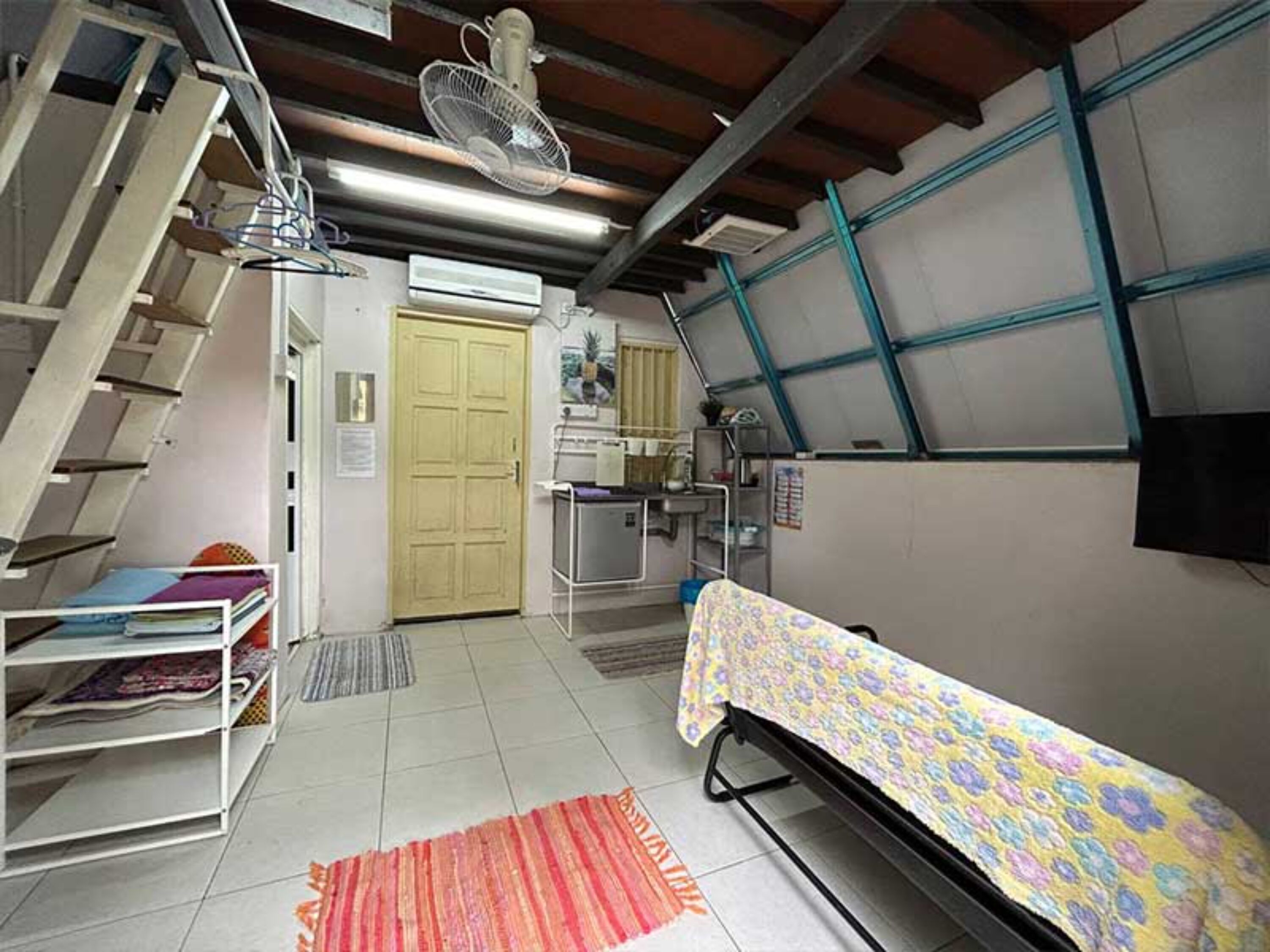 Une chambre avec un lit superposé, une petite table recouverte d’une nappe à motifs floraux, un ventilateur et un appareil de climatisation.