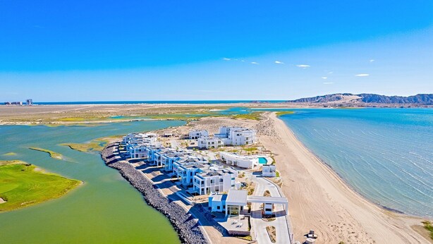 Un resort costero con edificios blancos modernos, una piscina y una playa.